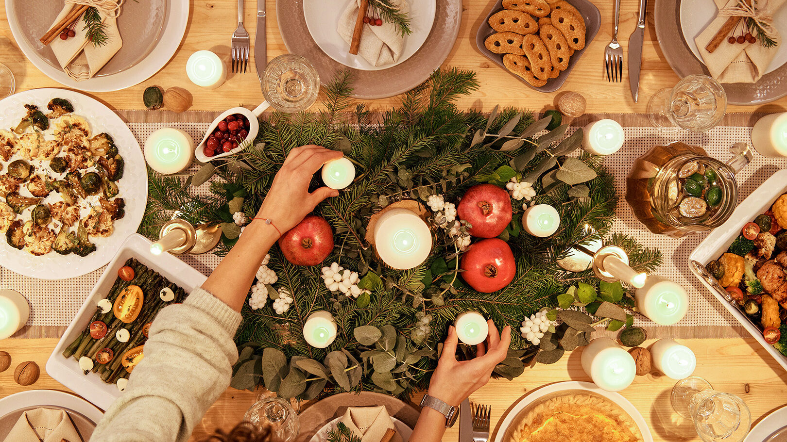 Weihnachtlich gedeckter Tisch mit Tannenschmuck und Essen, Frauenhände stellen gerade brennende Kerzen auf den Tisch