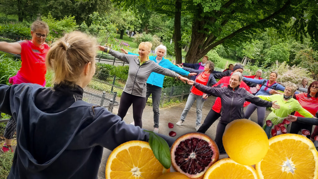 Eine Gruppen von Menschen bewegt sich zu Musik nach Anleitung einer Trainerin im Park. am Rand sind Früchte eingeblendet