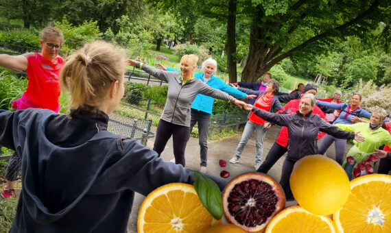 Eine Gruppen von Menschen bewegt sich zu Musik nach Anleitung einer Trainerin im Park. am Rand sind Früchte eingeblendet