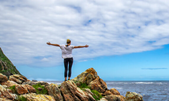 Frau mit ausgebreiteten Armen steht auf einem Felsen am Meer und sieht auf die Brandung