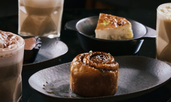 drei Kaffeegläser mit Milchschaum und zwei Stück Kuchen und Gebäck angerichtet auf dunklen Keramiktellern