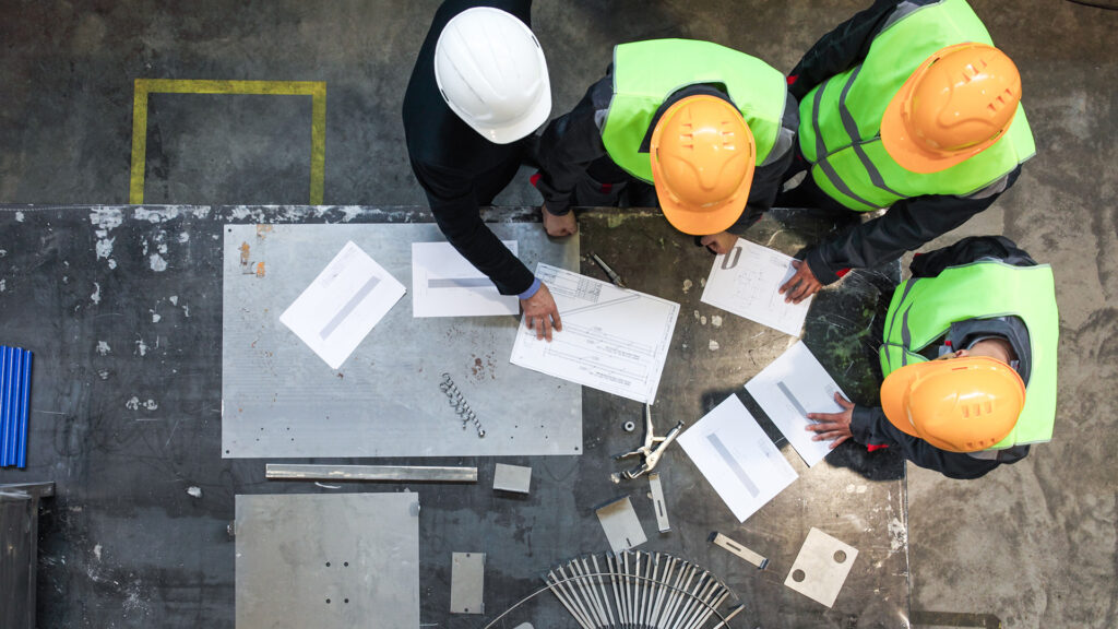 Arbeiter mit Schutzhelmen in der Industrie an einer Werkbank mit Werkzeugen und Metallteilen vor sich, besprechen sich