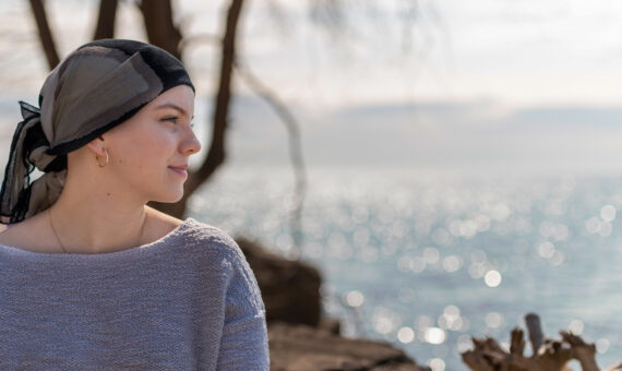 Eine junge krebskranke Frau lächelt selbstbewusst und schaut in die Ferne. Sie trägt ein Kopftuch und entspannt sich im Freien. Im Hintergrund sind ein See und ein Baum zu sehen. Es ist ein sonniger, friedlicher Tag.